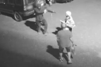A man points a shotgun at a woman and a man in a carpark in black and white CCTV vision.
