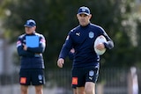 NSW State of Origin coach, Brad Fittler, holds a ball under his arm as he watches team training.