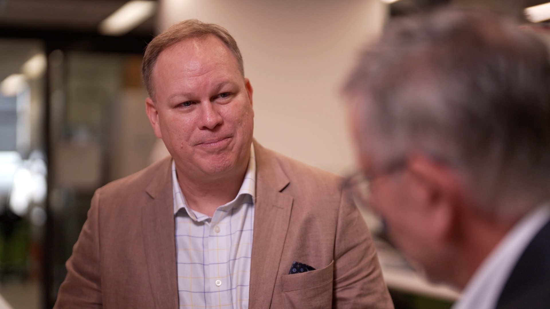 A man wearing a business shirt with a large check print and tan jacket talking to a man with grey hair and glasses.