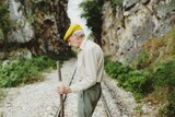 Elderly man with bright yellow flat cap stands on railway in gully