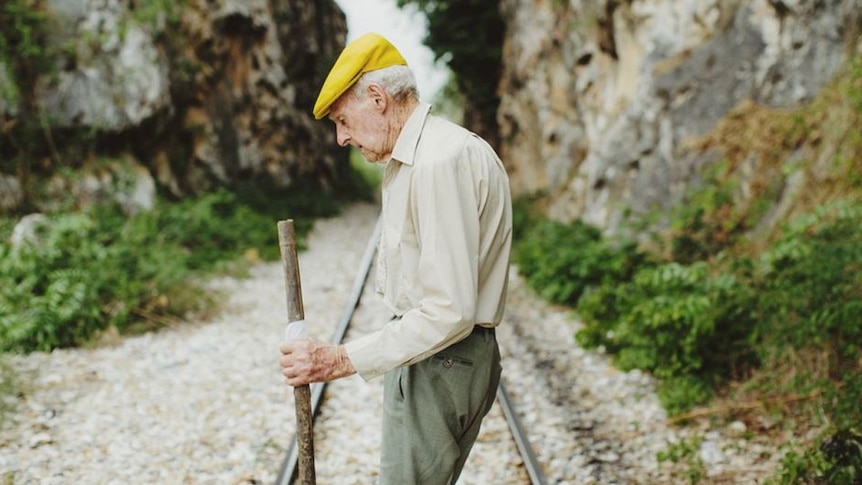 Elderly man with bright yellow flat cap stands on railway in gully