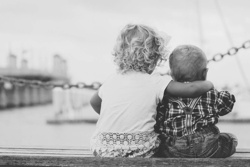 A black and white image shows two children sitting next to each other on a pier.