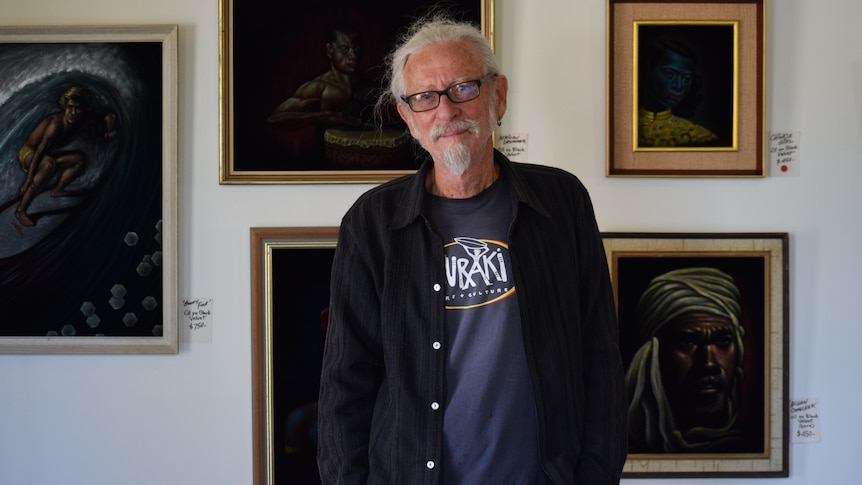 Man standing in front of black velvet paintings, smiling, wearing glasses. has long hair and beard