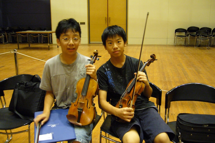 Brett Yang and Eddy Chen as young boys, sitting, holding violins.