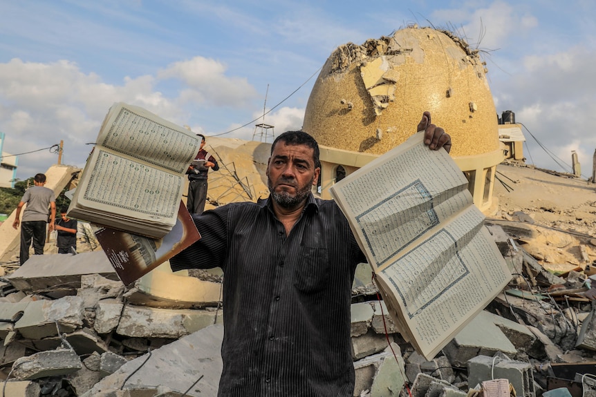A man holding books of the Quran in front of a destroyed mosque.