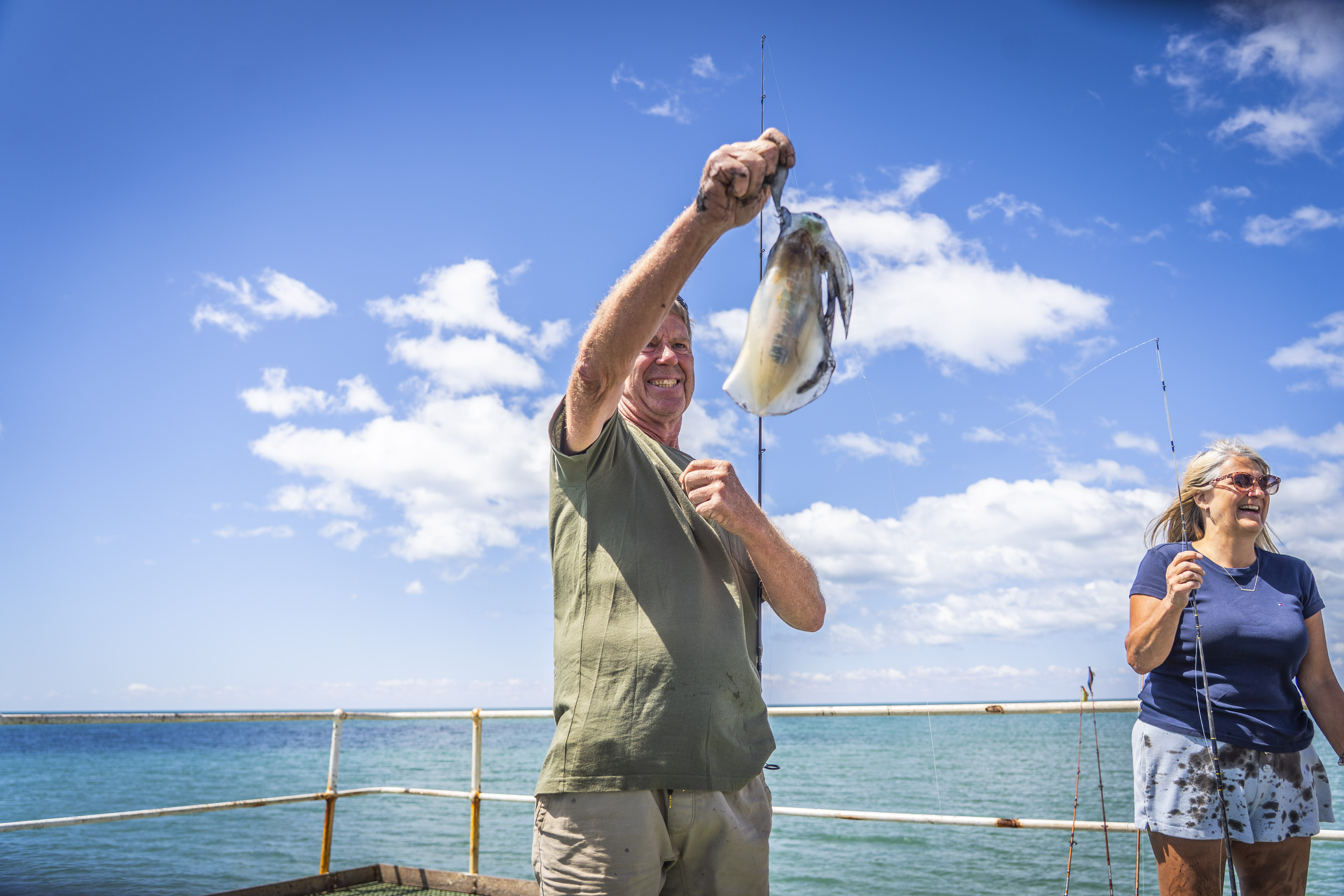 Um pescador tira o pescado da linha em um dia ensolarado, com o céu azul, nuvens brancas e o mar azul-turquesa atrás dele.