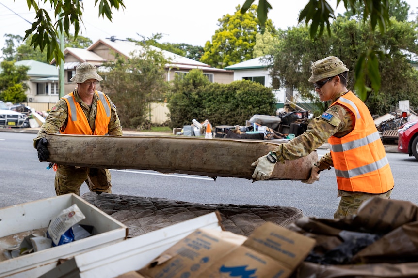 Two army members wearing high vis orange vests carry a mattress onto a pile of flood-damaged goods on a street