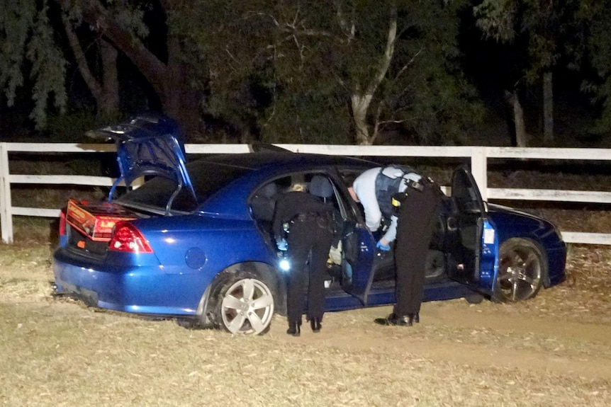 Two police officers peer inside a blue sedan with burst tyres.