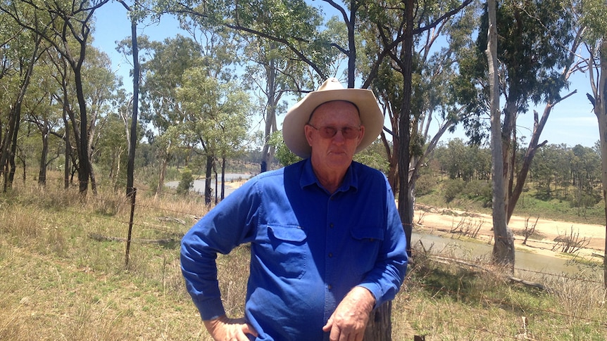 A man in his seventies leans against a fence wearing an Akubra hat with a river and trees in the background