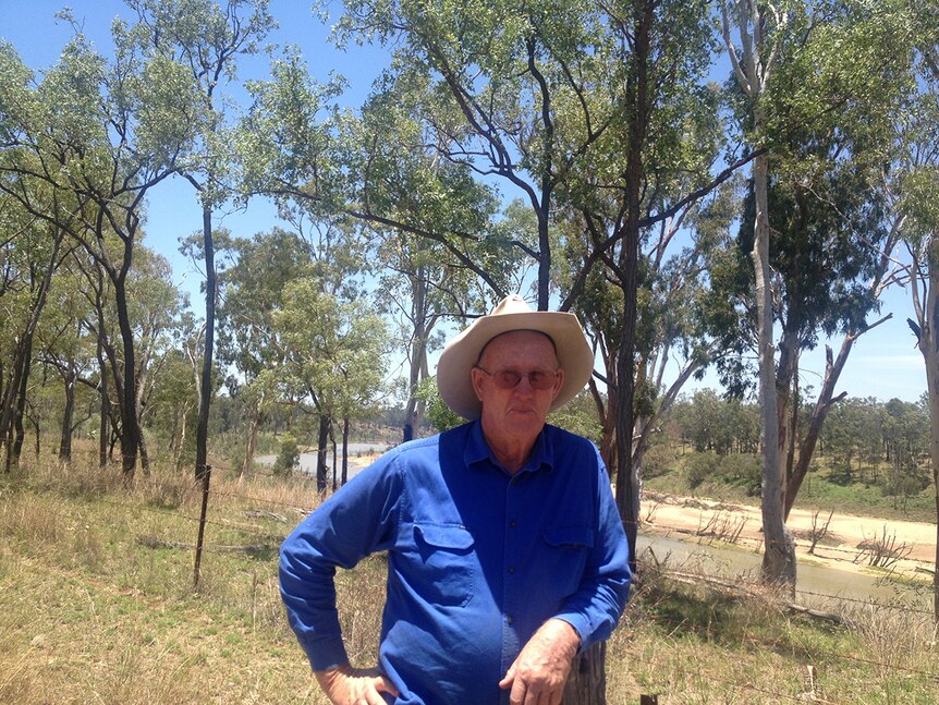 A man in his seventies leans against a fence wearing an Akubra hat with a river and trees in the background