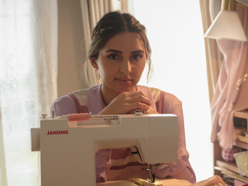 A young woman with strands of hair loosely failing around her face sits at a sewing machine. She looks serenely into the camera