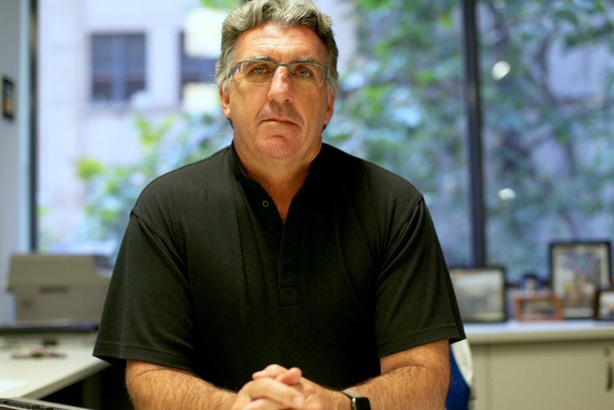 A serious middle-aged man wearing black shirt, grey hair, glasses looks at the camera, blurred glass windows behind.