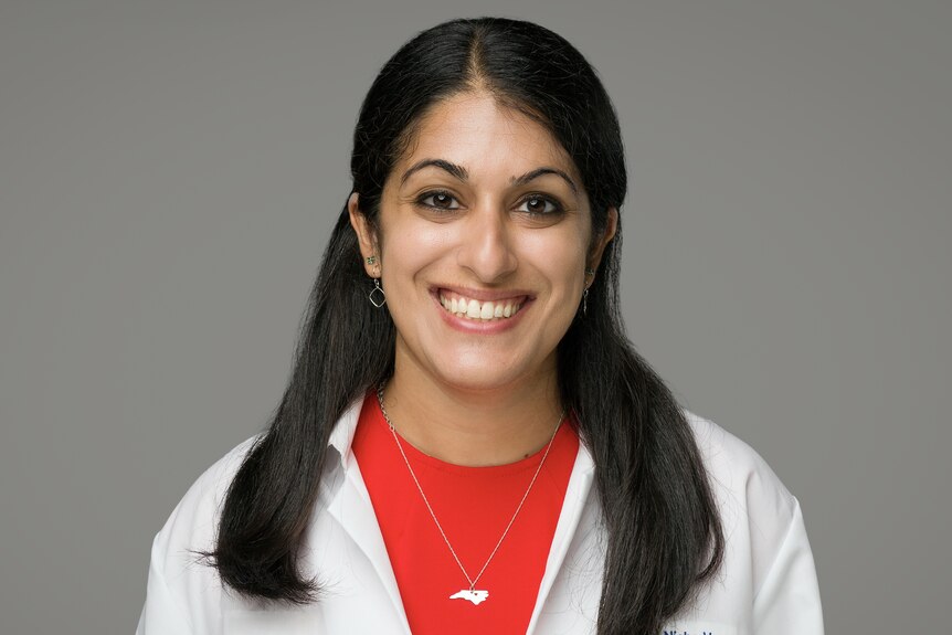 A woman wearing a white doctor's coat over a red top smiles to camera