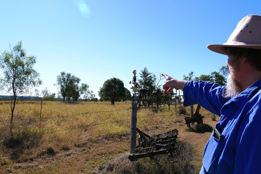 A man in a blue shirt and farming broad brim hat points over a fence.