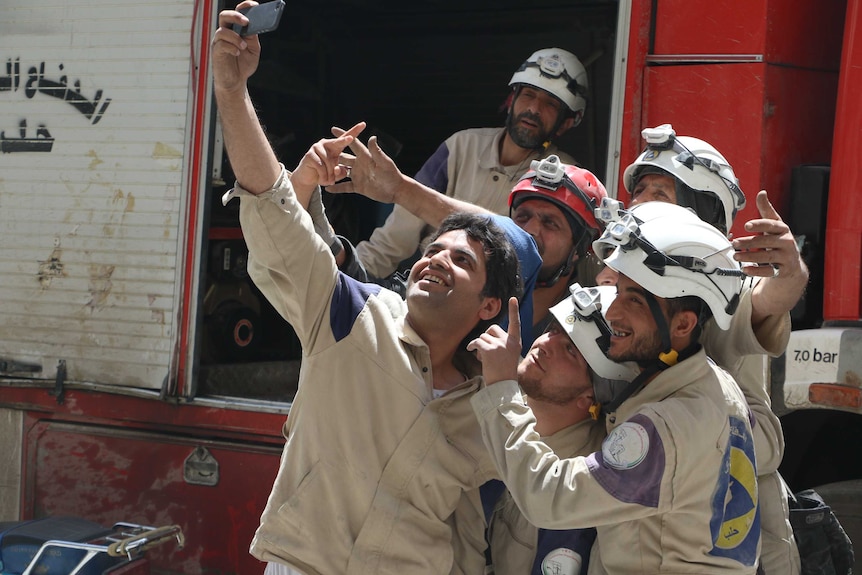 You view a crowd of men in white helmets and in beige work uniforms posting for a smartphone selfie.