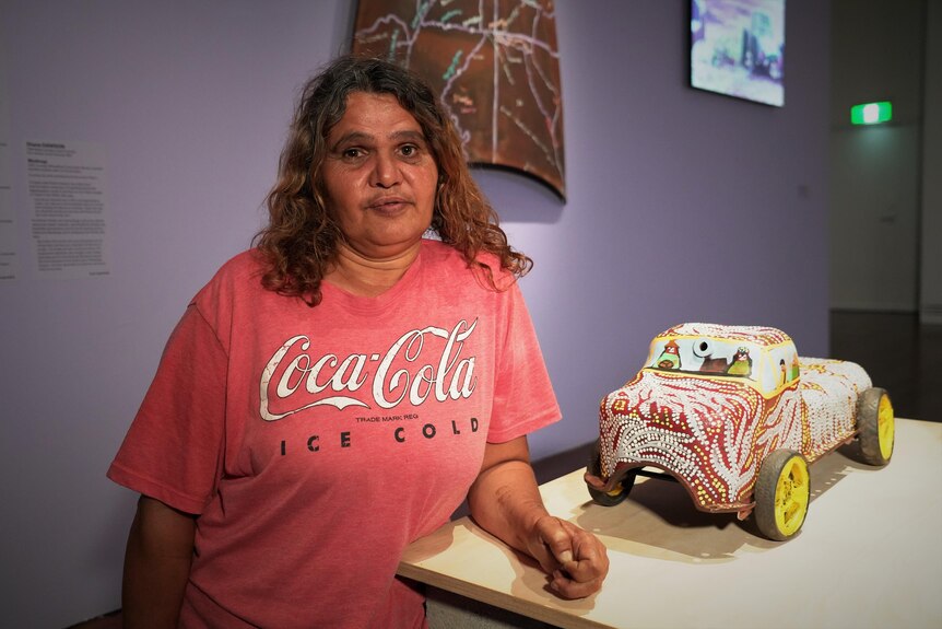 A woman with brown hair in a pale red Coca Cola t-shirt stands beside a painted sculpture of a car