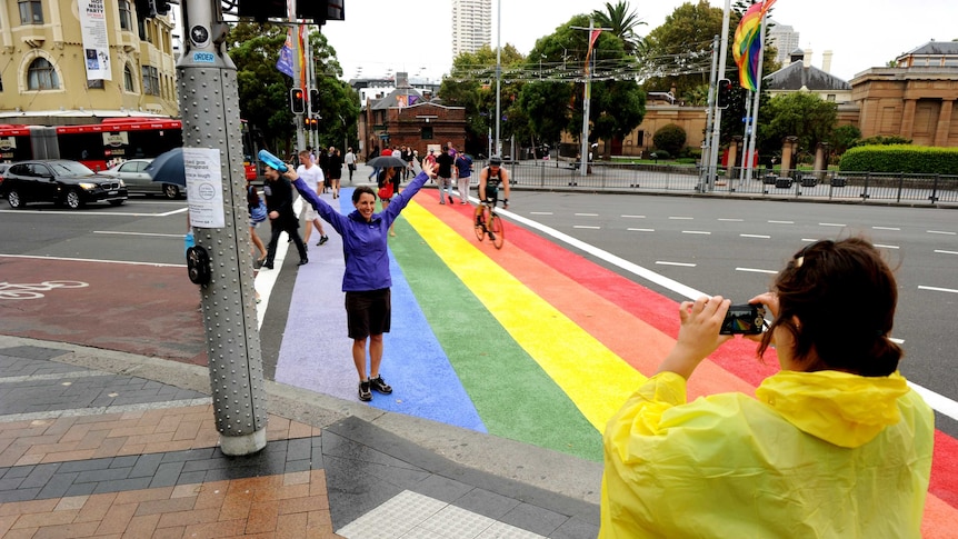A woman poses for a photo on the rainbow crossing while other pedestrians cross the road.