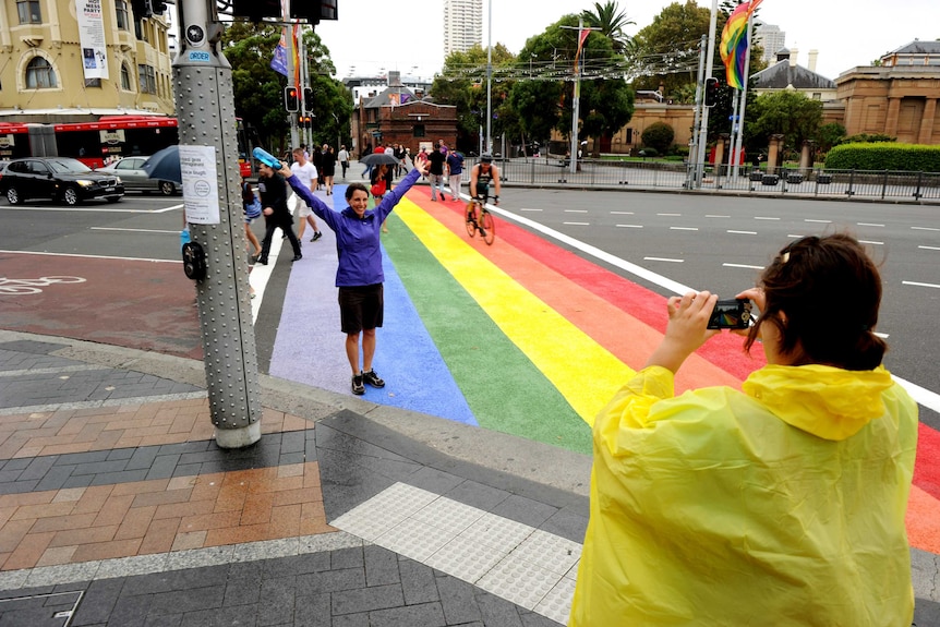 A woman poses for a photo on the rainbow crossing while other pedestrians cross the road.