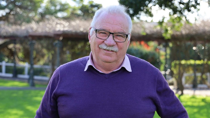 A moustached man wearing glasses and a purple jumper smiles at the camera while standing in a garden