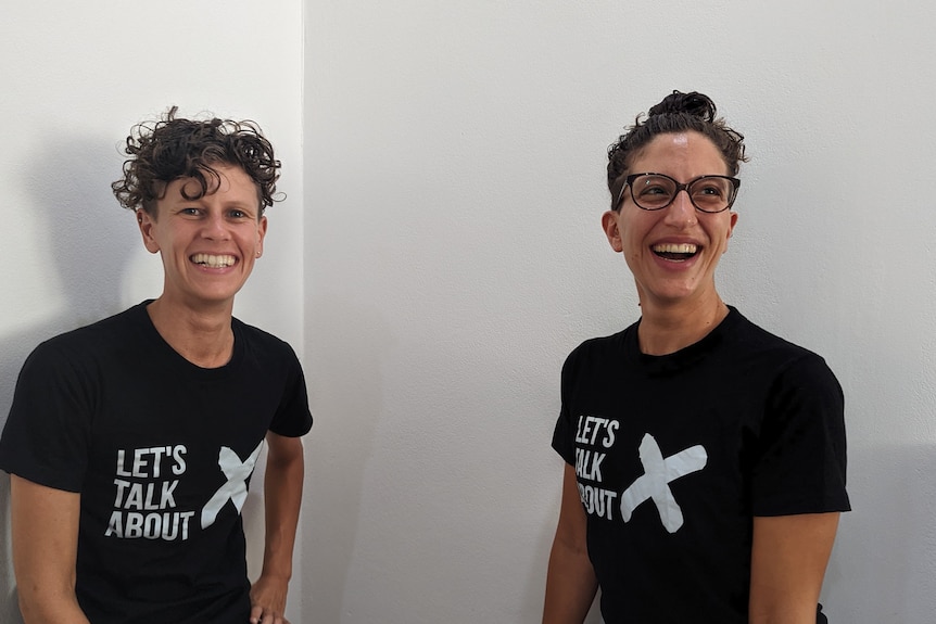 Two people with short dark hair smiling. They are both wearing dark T-shirts that says "Let's Talk About X".