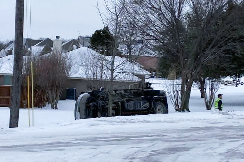A car lies on its side near a road as snow blankets the ground and trees.