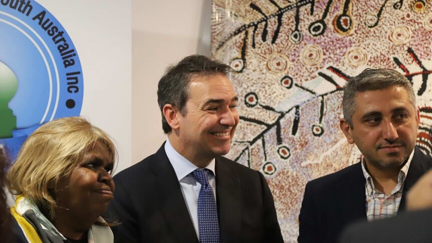 An aboriginal woman and two men smile and stand in front of an Aboriginal painting.