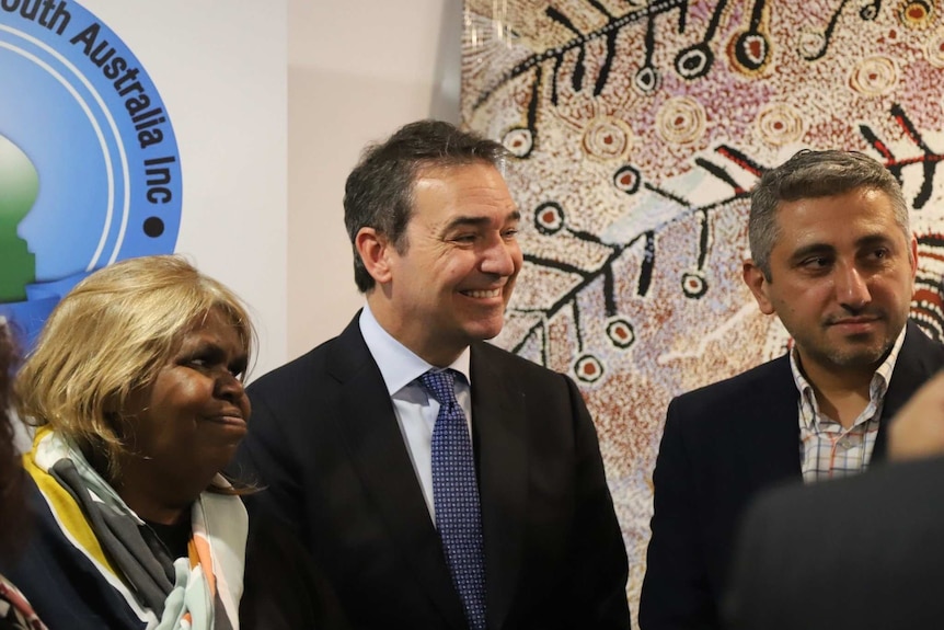 An aboriginal woman and two men smile and stand in front of an Aboriginal painting.