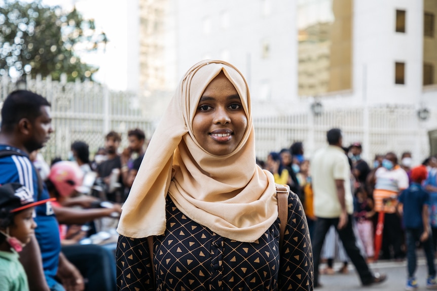 A young woman in a creamy coloured hijab smiles for the camera while standing on a busy street 