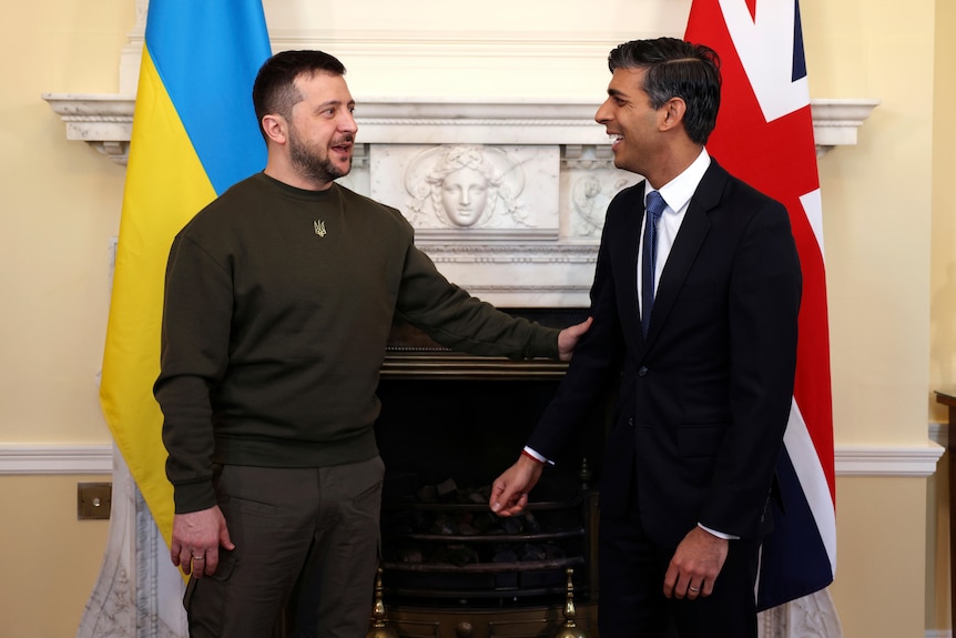 Zelenskyy reaches out to grasp Sunak's elbow as the pair smile at each other standing in front of their respective flags