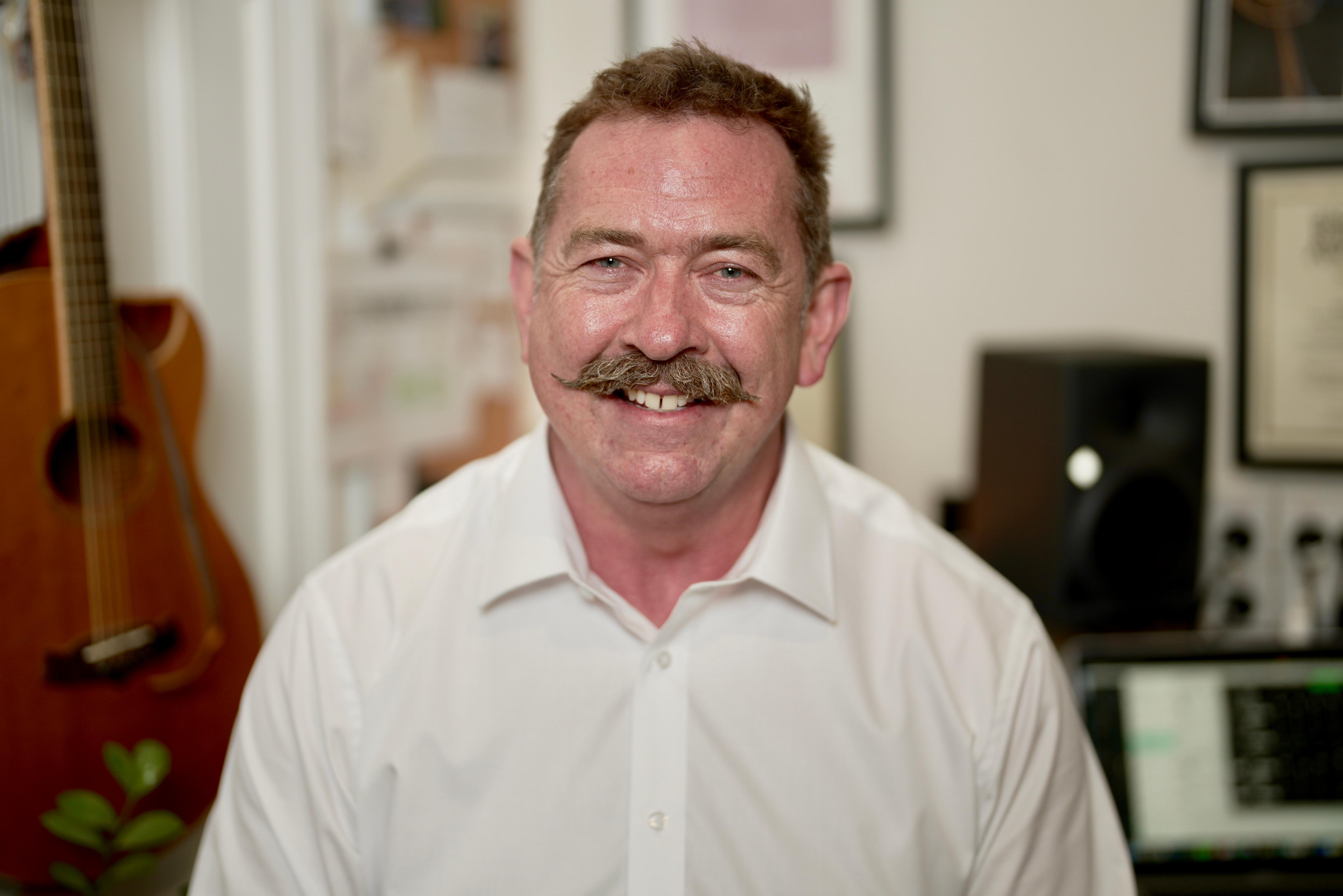 A man smiling with a guitar and trophies behind him. 