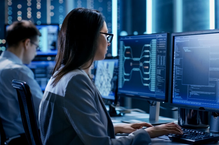 Two people use computers in a dark room lit with white and blue neon lights.