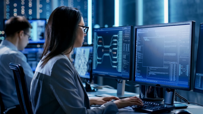 Two people use computers in a dark room lit with white and blue neon lights.