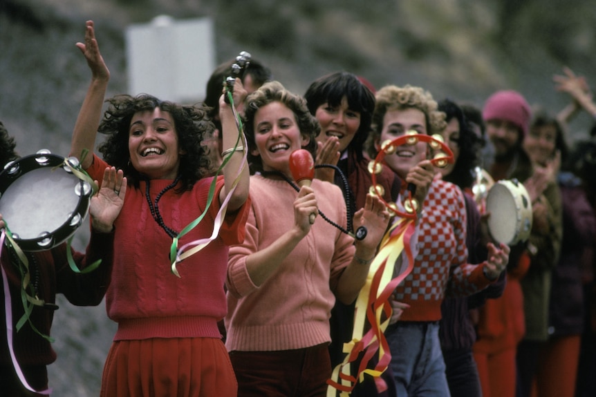 Women dressed in pink and orange in a line, dancing and playing musical instruments