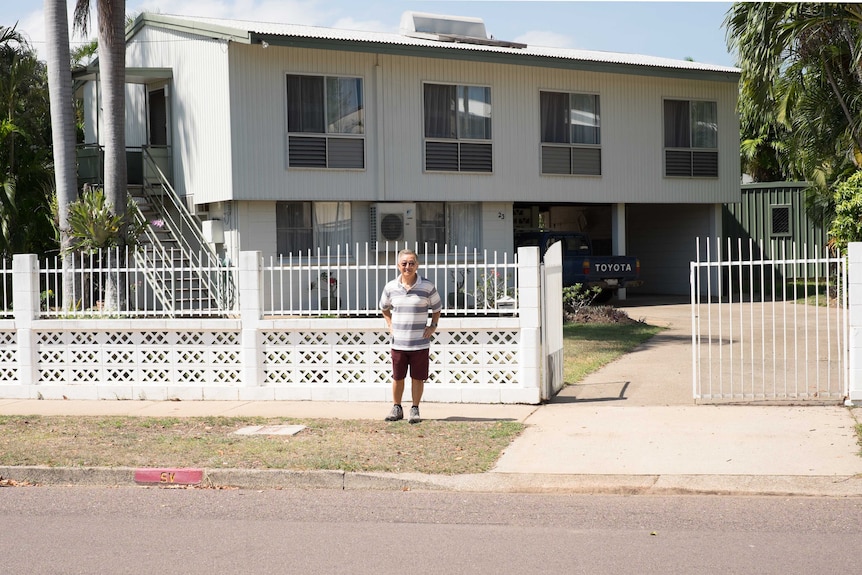 An elderly man stands in front of a house.