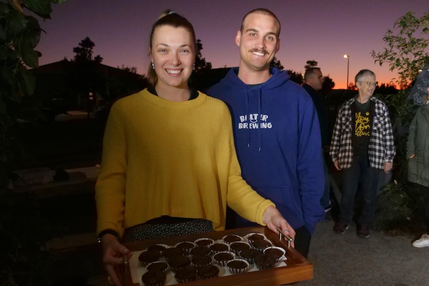 A smiling woman holding a tray of cupcakes stands next to her husband.