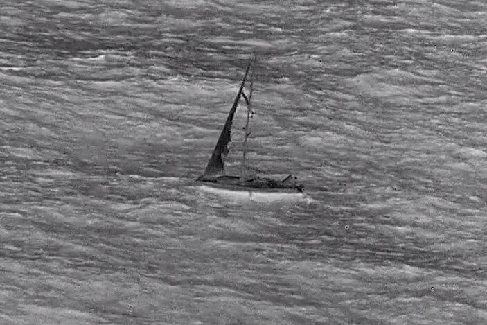 A grey-scale image of a sailing boat on the ocean, with apparent damage to the sail. 
