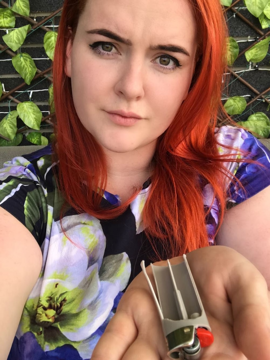 A young woman with red hair holds up the remains of an exploded lighter.