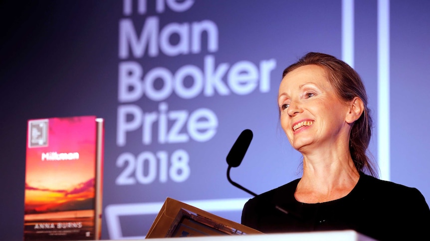 A woman smiles in front of projected text that reads "The Man Booker Prize 2018" and a copy of a book