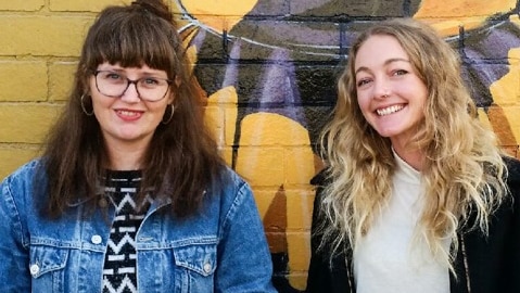 Two women standing in front of a yellow wall.