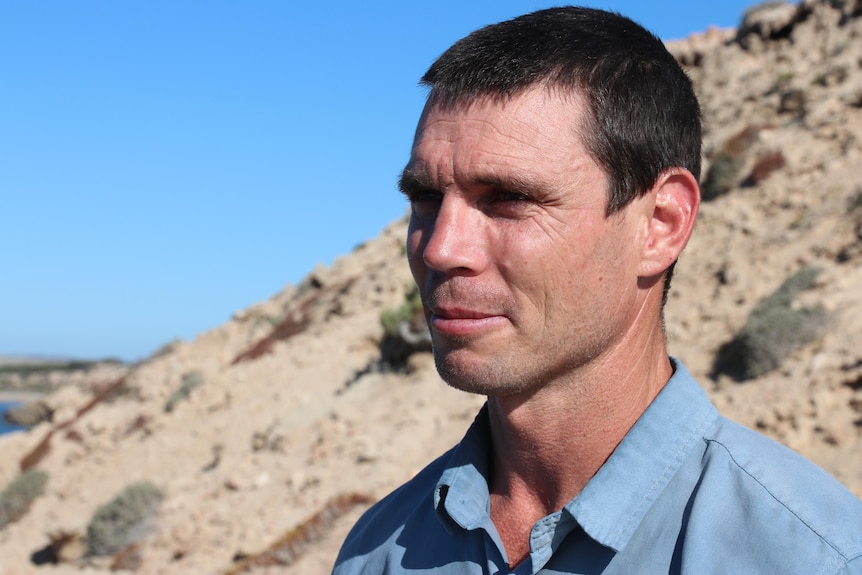 A close-up of a brunette man looking out to the horizon. He is wearing a blue shirt and stands in front of sandy dunes. 
