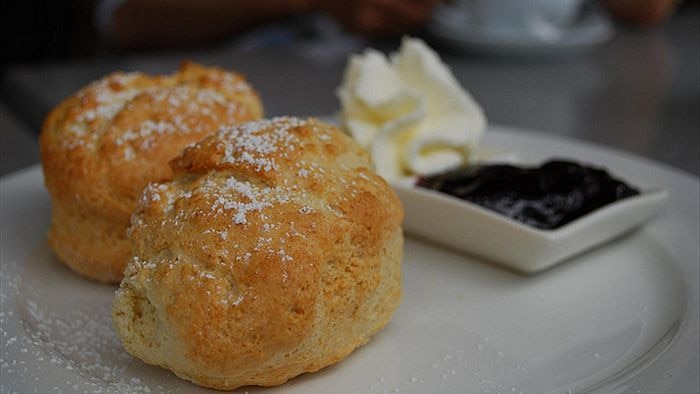 Two scones on a plate with a dish of jam and cream in the background.