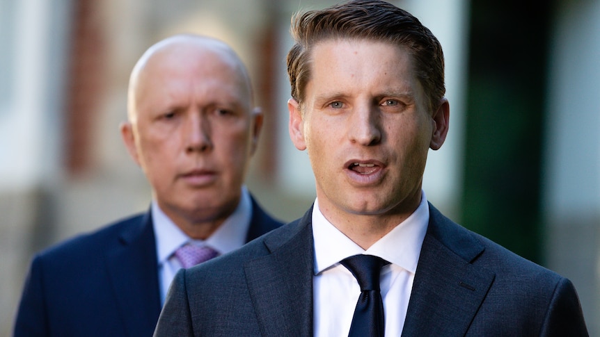 Andrew Hastie wears and suit and tie and is speaking with Peter Dutton watching in the background