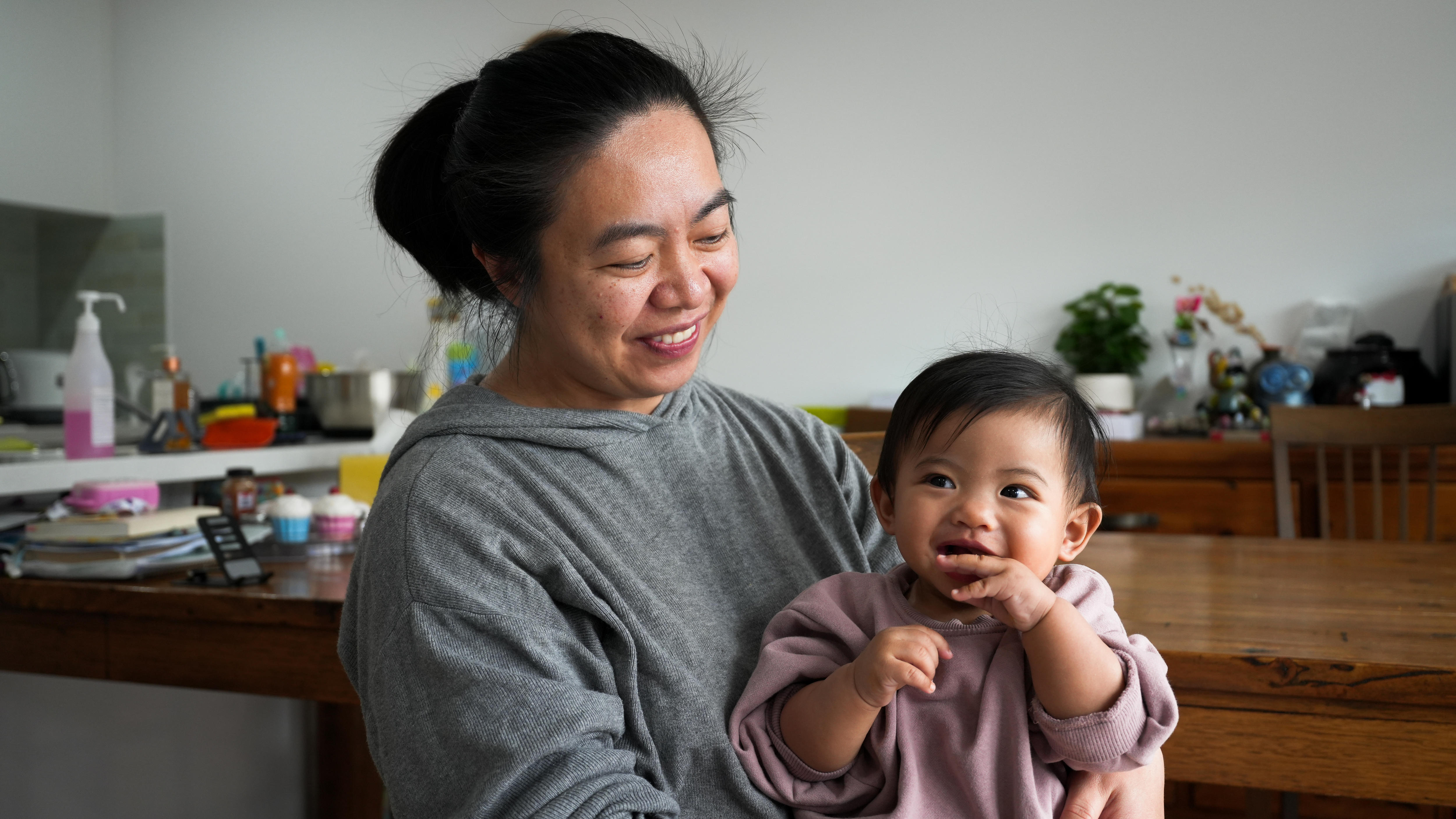 A woman wearing a grey jumper looking down smiling at a baby sitting in her lap