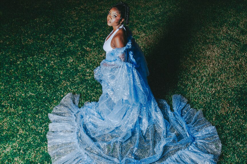 Chanel Loren, a Black woman, wearing a sprawling blue dress, gazing at the camera, surrounded by green grass