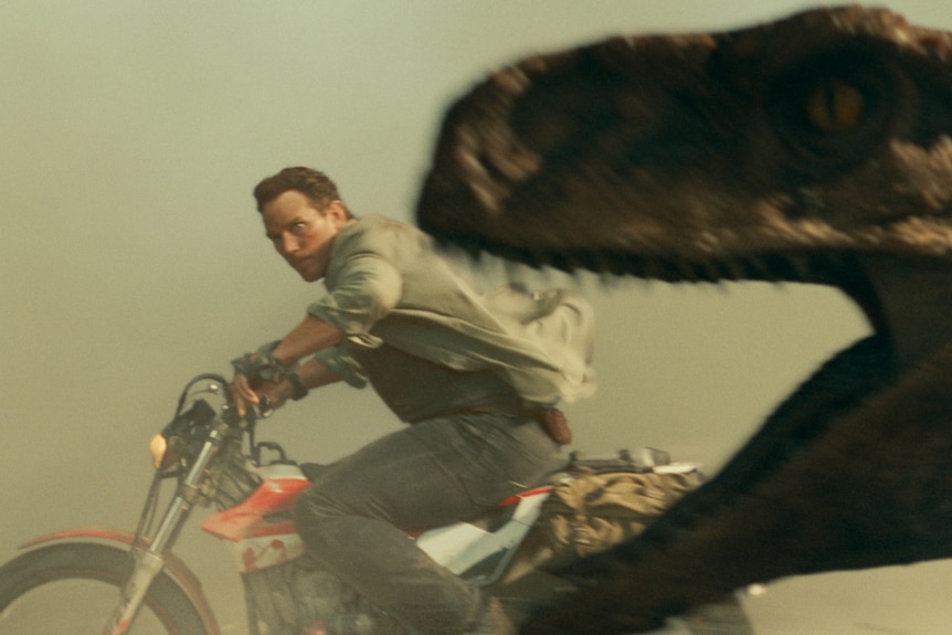 Un homme blanc aux cheveux bruns chevauche une moto à travers un panache de poussière, poursuivi par un vélociraptor.