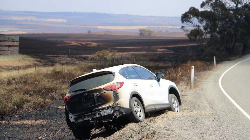 A partially burnt car sits by the side of the road, with burnt out landscape stretching into the distance.