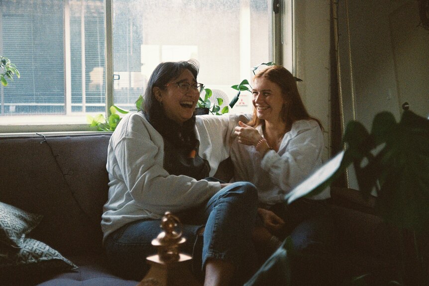 Two young women sit on the couch in a lounge room, laughing together.