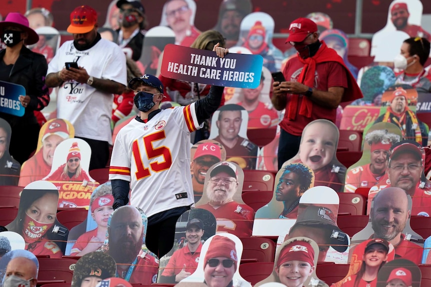 A Super Bowl fan holds up a signing reading "THANK YOU HEALTHCARE HEROES" while surrounded by cardboard cutouts of fans.