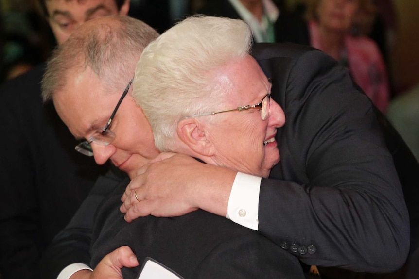 An elderly woman cries as Scott Morrison hugs her tightly. Her face is screwed up as she leans into his chest.