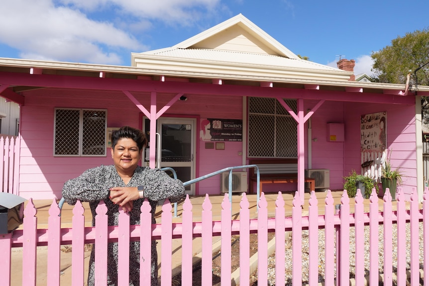 A woman leans on a pink fence in front of a pink house